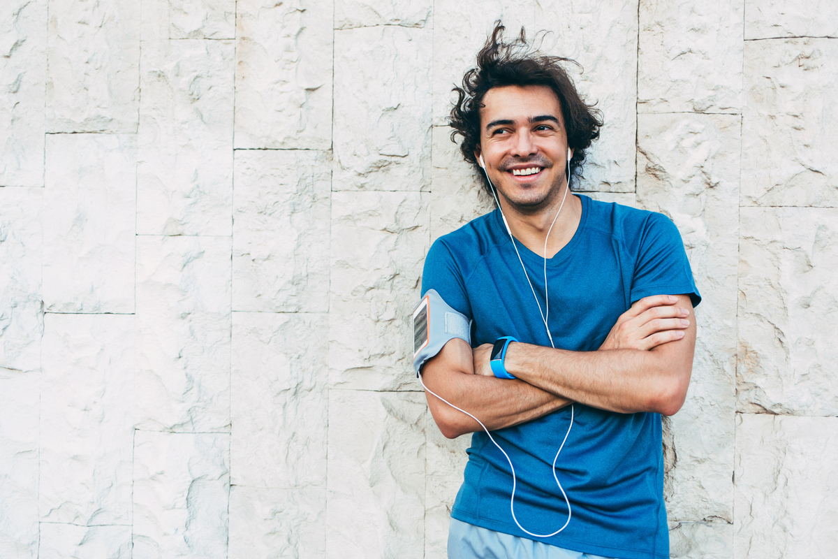 Smiling man in blue shirt leans against a white rock wall, looking to his right with his arms crossed and headphones on.